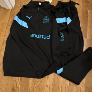 OM x Puma svarta träningsbyxor - Svarta träningsbyxor från Puma x Olympique de Marseille med blå detaljer och klubbmärke på låret. Byxorna har smal passform, elastisk midja med snörning och ventilerande meshpaneler. Perfekta för fotbollsträning eller chill.