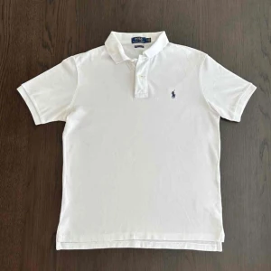Ralph Lauren pike - Klassisk vit pikétröja från Ralph Lauren med blå broderad logga på bröstet. Tröjan har krage, korta ärmar och knäppning framtill. Modellen är custom slim fit och tillverkad i bomull, perfekt nu i solen