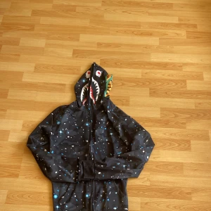 Svart hoodie med shark-hood från BAPE - Säljer en svart BAPE hoodie med ikonisk haj-hood och camo/galaxy-mönster. Dragkedja hela vägen upp över huvan, broderade detaljer och färgglada tryck på huvan. Perfekt statement-plagg för streetwear-älskare.
