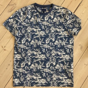 Blåvit mönstrad t-shirt från Gant - Snygg blå t-shirt från Gant med vitt naturinspirerat mönster över hela plagget. Klassisk rund halsringning och korta ärmar. Perfekt för dig som gillar en unik och stilren look. Tillverkad i mjuk bomull som känns skön mot huden.