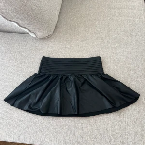 Svart skinn kjol - Snygg svart kortkjol från Gina Tricot i skinnimitation med bred ribbad midja. Kjolen har en lätt utställd form som ger en cool och trendig vibe. Perfekt för dig som vill sticka ut lite extra.