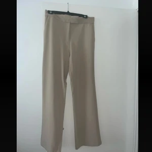 Beiga vida kostymbyxor - Stilrena beiga kostymbyxor med vida ben och hög midja. Byxorna har en klassisk look med dold hyskknäppning i midjan och är sydda i ett slätt, mjukt tyg som ger en dressad känsla. Perfekta för en clean och trendig outfit.