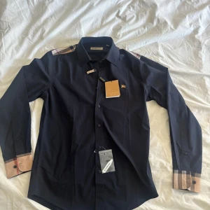Burberry Skjorta - Säljer denna stiliga Burberry Skjortan i storlek S. Aldrig använd och kommer med tags! Unik skjorta med burberry mönstret på baksidan också! Hör av dig vid funderingar! 