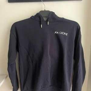 Svart hoodie Ma Chérie Gina Tricot - Svart hoodie från Gina Tricot med vit text 'MA CHÉRIE' och hjärtan tryckt både fram och bak. Tröjan har huva, snörning och långa ärmar. Mjuk insida och klassisk passform, perfekt för en chill look.