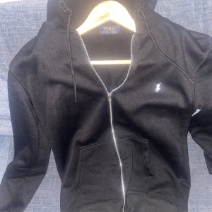 Svart hoodie från Polo Ralph Lauren - Svart hoodie från Polo Ralph Lauren med dragkedja, huva med snörning och klassisk logga broderad på bröstet. Tillverkad i 100% polyester och har praktiska fickor framtill. Perfekt för en chill och stilren look. Pris kan diskuteras!!