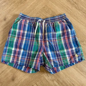 Rutig badshorts från Polo Ralph Lauren - Färgglada badshorts från Polo Ralph Lauren i rutigt mönster med blått, grönt, rött och vitt. Klassisk gul broderad logga, elastisk midja med snörning och innerbyxa i mesh. Perfekta för stranden eller poolen.