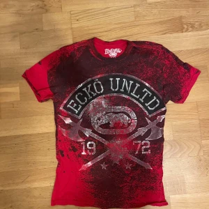  Ecko Unltd t-shirt - Cool röd t-shirt från Ecko Unltd med stora tryck både fram och bak, MMA-inspirerad design och svart-röda färgskiftningar. T-shirten har rund hals, korta ärmar och är gjord i 100% bomull. Perfekt för dig som gillar streetwear och sportig stil.