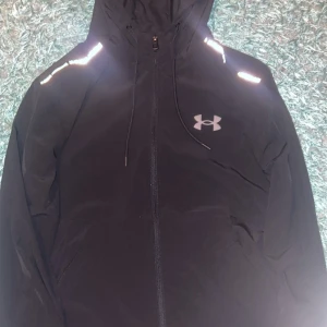 Svart vindjacka från Under Armour - Svart vindjacka från Under Armour med huva och reflekterande detaljer på axlarna. Jackan har dragkedja framtill, justerbar dragsko i huvan och logga på bröstet. Perfekt för dig som gillar sportig stil och vill synas i mörkret. Topp skick 10/10 inte använd endast prövad. M i VUXEN STORLEK lite slim modell 