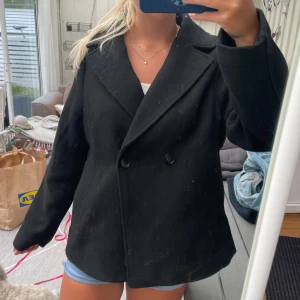 Svart skepparkavaj med knappar - Säljer en svart skepparkavaj med klassisk dubbelknäppning och bred krage. Jackan har en rak och loose passform, långa ärmar och är gjord i ett tjockare material som känns som ull. Perfekt för dig som gillar stilrena och tidlösa plagg.