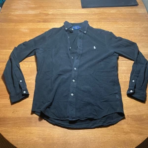 Svart skjorta från Ralph Lauren M - Svart långärmad skjorta från Ralph Lauren i bomullsmaterial, med klassisk krage och vita knappar framtill. Skjortan har den ikoniska broderade loggan på bröstet och är i modellen Featherweight Mesh, vilket gör den extra lätt och skön att bära.