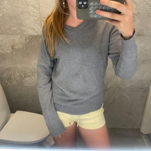 Grå v-ringad tröja Tommy Hilfiger - Snygg grå v-ringad tröja från Tommy Hilfiger i pima bomull och kashmir. Klassisk modell med liten broderad logga på bröstet och ribbade muddar. Perfekt till jeans eller kjol för en clean och stilren look.