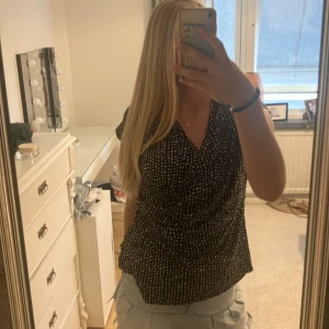 Svartvit omlottblus från Indiska - Svartvit omlottblus från Indiska i storlek XL. Blusen har korta ärmar, v-ringning och ett prickigt mönster. Lätt och luftig känsla, perfekt till jeans eller kjol. Snygg omlott-effekt framtill som ger en schysst siluett.