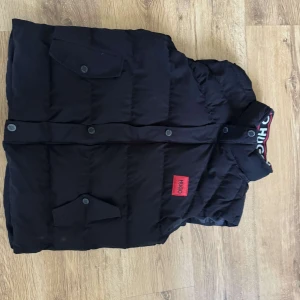 Svart dunväst från Hugo Boss - Svart dunväst från Hugo Boss med röd Hugo-logga på bröstet och tryckknappar med märkets namn. Västen har två fickor med lock och en hög krage med snyggt Hugo-tryck på insidan. Perfekt för lager-på-lager och streetstyle.