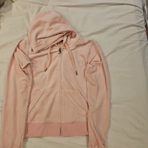 Rosa velour hoodie från Juicy Couture XS - Supermjuk rosa hoodie från Juicy Couture i velour med dragkedja och huva. Klassisk JC-logga broderad på bröstet och snygg J-detalj på dragkedjan. Perfekt för en chill och trendig look.