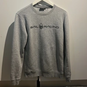 Grå Sail Racing sweatshirt - Snygg grå sweatshirt från Sail Racing med svart logga tryckt på bröstet. Klassisk rund halsringning och långa ärmar. Perfekt basictröja med stilren look, passar till jeans eller joggers. Mjuk och skön bomullsblandning.