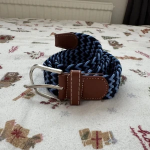 Blått flätat bälte med läderdetaljer - Snyggt flätat bälte i två nyanser av blått med bruna läderdetaljer och silvrigt spänne. Bältet har ett coolt zigzag-mönster och passar perfekt till jeans eller chinos för en avslappnad men stilren look. (Helt ny). 
