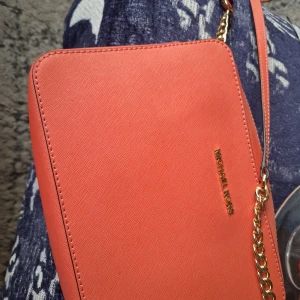 Orange axelväska från Michael Kors - Snygg orange axelväska från Michael Kors med guldfärgad kedjedetalj på axelremmen. Väskan är i slätt skinn och har en stilren, rektangulär form med diskret logga i guld på framsidan. Perfekt för att lyfta vilken outfit som helst.