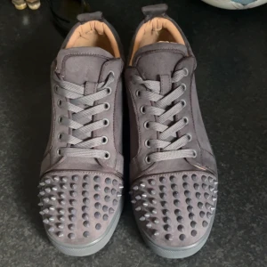Gråa Louboutin sneakers med nitar - Säljer ett par gråa sneakers från Louboutin med ikonisk röd sula och coola nitar på tån. Skorna är i mocka och har snörning framtill. Perfekta för dig som vill sticka ut med en edgy vibe och exklusiv känsla. Inget og ingår 