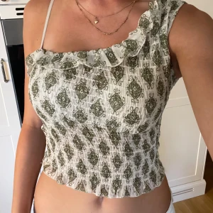Grönmönstrad one shoulder topp - Supersöt topp med one shoulder och volangkant. Tunt, smockat tyg i vitt med grönt mönster. Sitter tight och har en croppad passform. Perfekt för varma dagar och ger en trendig vibe med sin unika axel och volangdetalj. Köpt utomlands, priset kan diskuteras💕