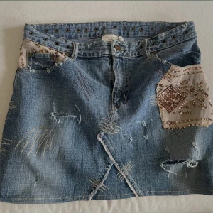 Jeanskjol med broderi från Karen Millen - Unik kort jeanskjol från Karen Millen med coola slitningar, broderade detaljer och inslag av beige tyg med pärlor och paljetter. Kjolen har nitar i midjan och en omlottliknande front. Perfekt för dig som vill sticka ut med en edgy och kreativ stil.