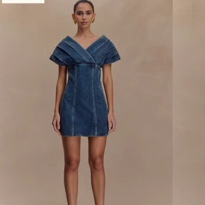 Kort jeansklänning med v-ringning - Säljer en snygg kort jeansklänning med markerad v-ringning och breda axelpartier. Klänningen har figurnära passform och coola sömdetaljer framtill. Perfekt för dig som vill sticka ut med en modern denimlook.