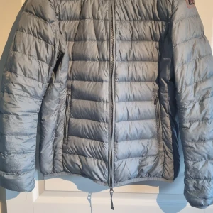 Ljusgrå/blå dunjacka Super Lightweight L - Ljusgrå/blå dunjacka i storlek L från Super Lightweight. Jackan är quiltad med horisontella sömmar, har dragkedja framtill och två sidofickor med dragkedja. Yttermaterialet är polyamid och fyllningen består av 90% dun och 10% fjädrar. Perfekt för kyliga dagar.