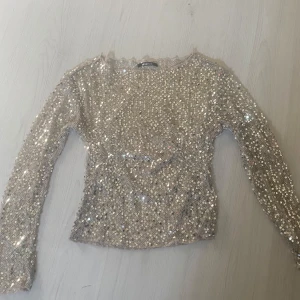 Glittrig paljettopp från Gina Tricot - Superglittrig långärmad topp från Gina Tricot, täckt med silvriga paljetter på ett nätliknande tyg. Toppen har rund halsringning och figurnära passform, perfekt för att sticka ut. Materialet är syntet och toppen är i en snygg ljus silverton.