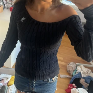 Mörkblå offshoulder stickad tröja Abercrombie - Snygg mörkblå stickad tröja från Abercrombie & Fitch med offshoulder-modell och kabelstickat mönster. Tröjan har långa ärmar och en vit älg-logga på bröstet. Perfekt för dig som vill ha en trendig och bekväm look.