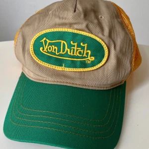Vintage Von Dutch grön, beige, gul keps, storlek ONE SIZE - Sällsynt keps från Von Dutchs originalproduktion på 1990-talet. Tillverkad i Korea i klassisk truckermodell med bomull och nylon – ett äkta Y2K-statement.  Skick: Bra vintage-skick utan fläckar, hål eller skador. Se bilder för egen bedömning.  Storlek: One size – justerbar passform (snapback)  Material: Bomull och nylon