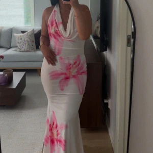 Vit långklänning med rosa blommor - Supersnygg vit långklänning med stora rosa blommor och djup urringning. Klänningen har öppen rygg och figurnära passform som framhäver siluetten. Perfekt för sommarens alla tillfällen när du vill sticka ut.