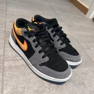 Nike Air Jordan 1 Low grå/orange - Snygga Nike Air Jordan 1 Low med grå mockadetaljer, svart textil och gula inslag på häl och swoosh. Skorna har svarta skosnören, vit sula och klassisk Air Jordan-logga bak. 