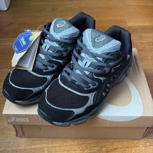 Svarta och grå Asics sneakers - Snygga Asics sneakers i svart och grått med meshpaneler och detaljer i syntet. Skorna har Ortholite innersula för extra komfort och GEL-dämpning i sulan. Perfekta för dig som gillar sportig stil och vill ha riktigt sköna skor till vardagen.