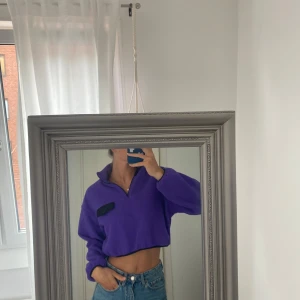 Lila croppad fleece half zip-tröja - Säljer en lila croppad fleece-tröja med half zip och hög krage. Tröjan har en svart knappdetalj på bröstet och elastisk nederkant. Perfekt för dig som gillar en chill och trendig stil. Passformen är loose och ärmarna är långa.