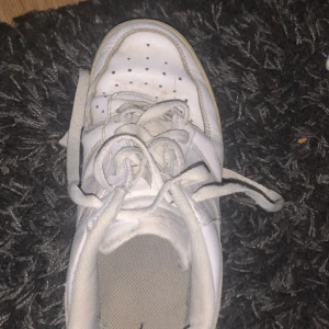 Vita Nike Air Force 1 sneakers - Klassiska vita Nike Air Force 1 sneakers i skinn med perforerad tå, snörning och ikonisk swoosh på sidan. Låg modell med platt sula och vadderad kant för extra komfort. Perfekta för dig som gillar streetstyle och tidlös design.