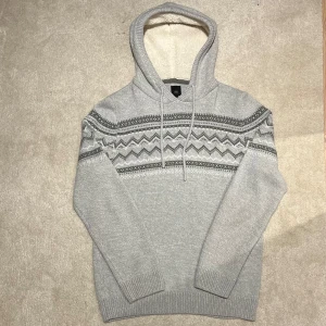 Grå stickad hoodie med mönster - Säljer en mysig grå stickad hoodie med ett snyggt mönster i mörkare grå nyanser. Hoodien har en fodrad huva och ribbade muddar vid ärmslut och nederkant. Perfekt nu till våren! Vid minsta fråga eller fundering hör av er🤝