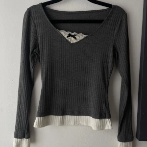 Ribbad grå långärmad tröja med spets - Mörkgrå ribbad långärmad tröja med v-ringning, vit spetskant och liten svart rosett vid halsen. 