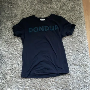 Mörkblå t-shirt från Dondup - Mörkblå t-shirt från Dondup med stort tryck över bröstet i ljusblått. Klassisk rund halsringning och korta ärmar. Tillverkad i mjukt material som känns skönt mot huden. Perfekt för en clean och stilren look. STORLEK S MEN PASSAR XS
