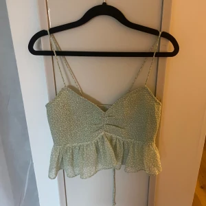 Ljusgrön croppad linnetopp med volang - Superfin ljusgrön croppad linnetopp med smala axelband och volang nertill. Toppen har ett diskret mönster, rynkdetalj framtill och knytning i ryggen. Perfekt för varma dagar och ger en söt och somrig vibe.