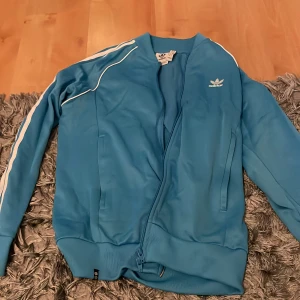 Blå Adidas bomberjacka med vita ränder - Snygg blå bomberjacka från Adidas med klassiska vita ränder längs ärmarna och vit logga på bröstet. Jackan har dragkedja framtill, ribbade muddar och är tillverkad i ett mjukt syntetmaterial. Perfekt för en sportig och avslappnad stil.