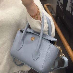 Ljusblå axelväska  - Snygg ljusblå axelväska i skinn med guldfärgade detaljer och ett justerbart axelband. Väskan har två handtag och stängs med en knapp framtill. Väskan är köpt på Raglady och är en av dom nyare märkena Latalia dom fått in.