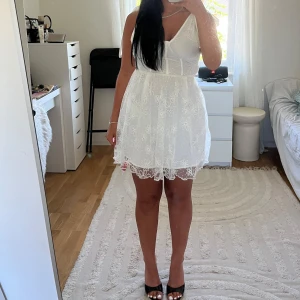 Vit kort spetsklänning med rosett - Supersöt vit kort klänning med broderade blommor och spetsdetaljer. Klänningen har v-ringning, axelbandslös look med stora rosetter på axlarna och även knyt vid ryggen. Den är även i helt nytt skick och med prislapp.