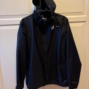 CDG vindjacka - Svart vindjacka från K-Way och Comme des Garçons PLAY med huva, dragkedja och ikoniska hjärtloggan på bröstet. Jackan är i lätt och smidig polyester, perfekt för blåsiga dagar. Snygg och minimalistisk design med coola detaljer och loggor framtill. Kvitto finns, storlek Medium, passar mellan 175-185