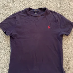 Mörkblå t-shirt från Polo Ralph Lauren - Snygg mörkblå t-shirt från Polo Ralph Lauren med klassisk röd broderad logga på bröstet. Dåligt ljus på bilden. T-shirten är i mjuk bomull och har en normal passform med korta ärmar och rund hals. Perfekt för en clean och stilren look. Kom med frågor och förslag! 