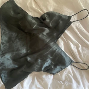 Svart satin linnetopp med tunna band - Snygg svart linnetopp i satin med tunna axelband och glansig finish. Toppen har en lös passform och spetsig nederkant, perfekt för att styla med jeans eller kjol. Enkel men ändå trendig look.