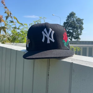 New era keps  - Snygg svart New Era 59FIFTY Yankees keps med vit NY-logga framtill, broderade röda rosor på sidan och World Series-märke. Klassisk flat peak och broderade detaljer. Tillverkad i polyester för en clean och modern look. Fick kepsen i present men kända att den inte var min stil. Stlk 7 1/2 = 59,6