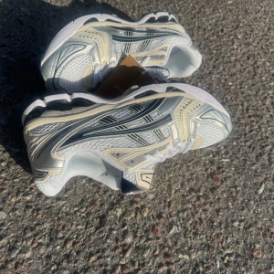 Vita och beige sneakers från Asics - Snygga Asics sneakers i vitt och beige med svarta detaljer. Ovandelen är i mesh och syntet för bra andningsförmåga och komfort. Klassisk snörning och sportig design med rund tå och platt sula. Perfekta för dig som gillar retrovibbar och streetstyle.