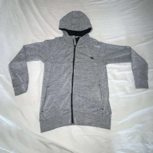 Grå hoodiejacka med dragkedja - Snygg grå hoodiejacka från CLN Athletics med hel dragkedja och två fickor framtill. Jackan har en sportig look, huva och svart logga på bröstet. Perfekt för chill eller träning, tillverkad i mjukt material som känns skönt mot huden.