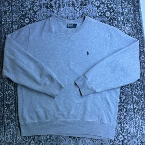 Grå sweatshirt från Polo Ralph Lauren - Klassisk grå sweatshirt från Polo Ralph Lauren med broderad marinblå logga på bröstet. Tröjan har rund hals, ribbade muddar och är långärmad. Perfekt basic-plagg med clean look och skön passform.