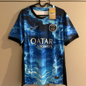 PSG x Nike blå fotbollströja - Snygg PSG fotbollströja från Nike med ett unikt blått moln- och stjärnmönster. Tröjan har korta ärmar, svart Nike-logga och Paris Saint-Germain-emblem på bröstet. Qatar Airways som sponsor framtill. Tillverkad i lätt och ventilerande material.
