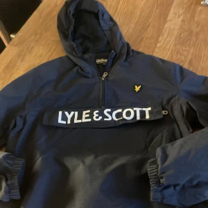 Blå vindjacka från Lyle & Scott - Snygg marinblå vindjacka från Lyle & Scott med huva och halv dragkedja. Jackan har stor logga framtill och klassisk gul fågel på bröstet. Perfekt för blåsiga dagar och enkel att slänga på sig över en hoodie.